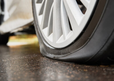 Stopped white car with punctured tire on roadside.