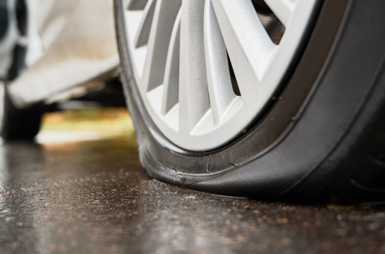Stopped white car with punctured tire on roadside.